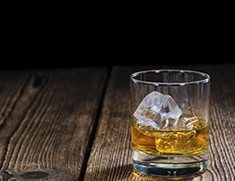 A tumbler of alcohol with ice sits on a wooden table.