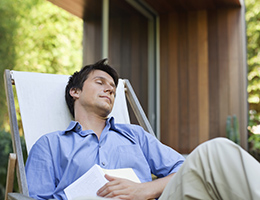 A middle-aged man in a blue shirt and tan pants is napping in an outdoor chair with a white cover. An open book rests on his stomach.